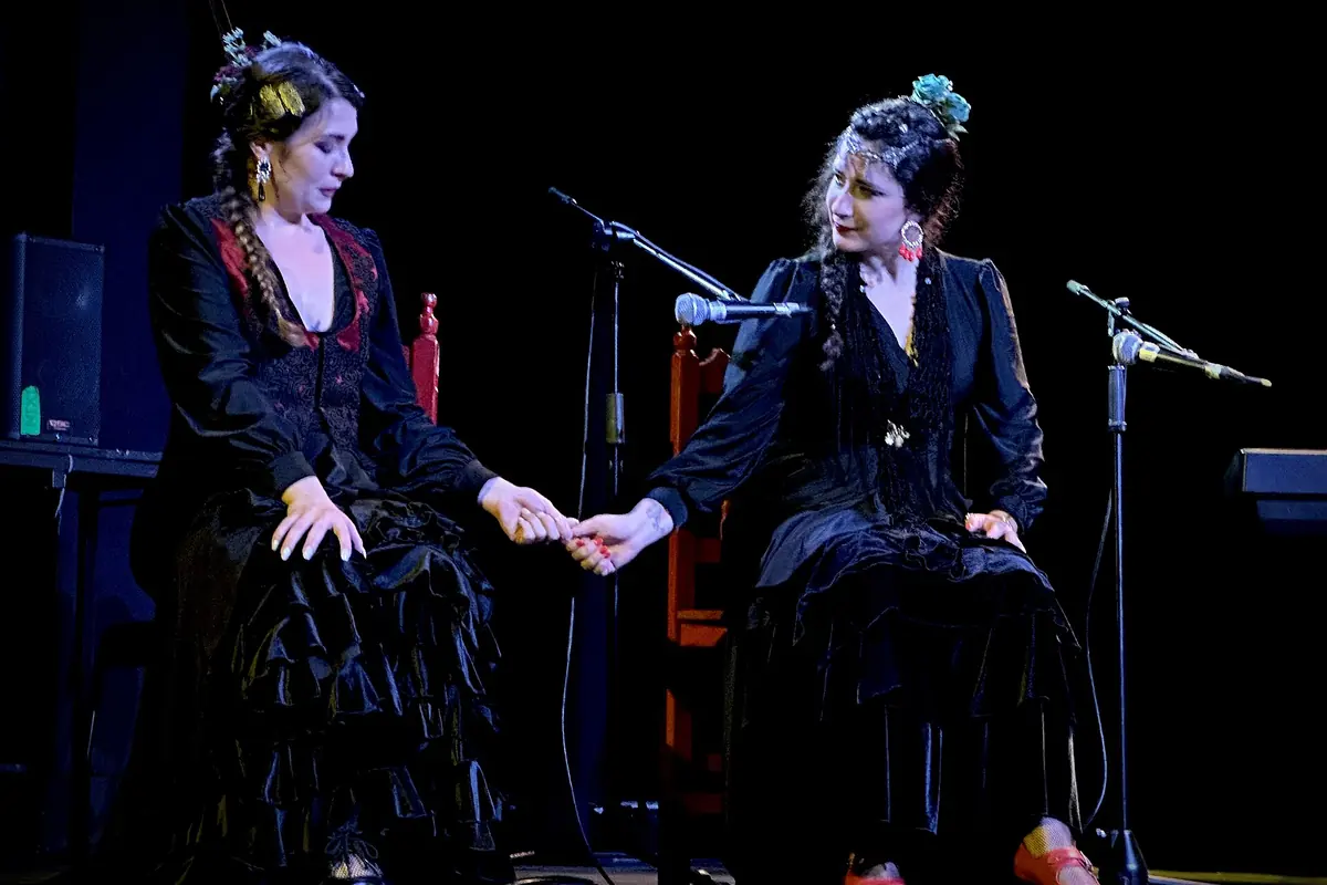 Two women in black dresses with flowers in their hair sit holding hands on a dark stage beside microphones