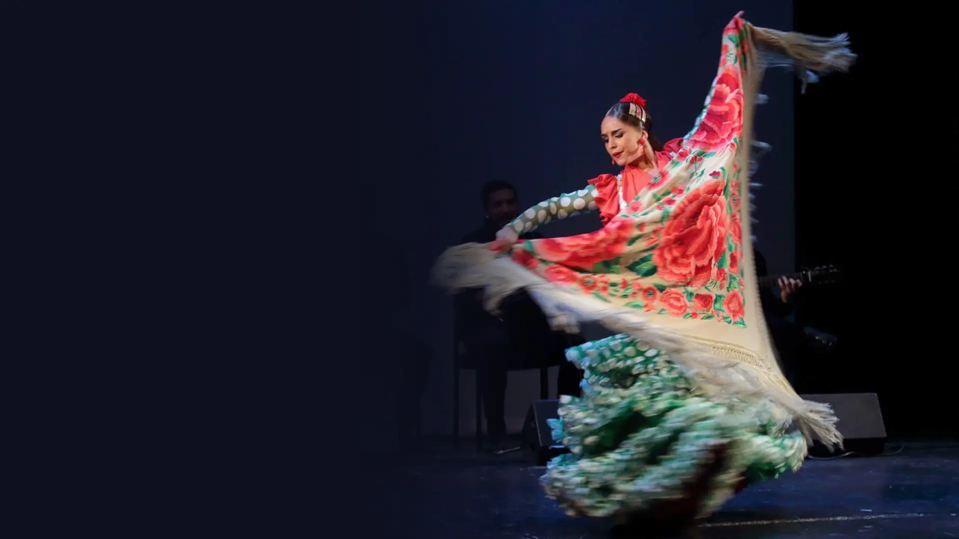 A flamenco dancer in a green polka-dot ruffled dress swirls a large cream shawl embroidered with red roses on a dark stage, with musicians seated behind her