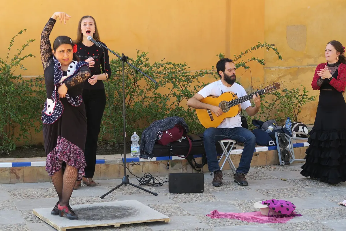 A dancer with one arm raised, a singer at a microphone and a seated guitarist perform in a courtyard with an ochre wall, while another dancer in a black ruffled dress claps