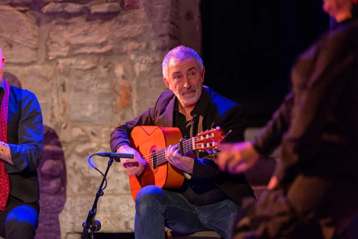 A grey-haired flamenco guitarist plays a golden guitar into a microphone while a dancer's arm moves in the foreground